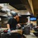 Japanese restaurant kitchen with chefs wearing black uniforms and hats preparing dishes. Diners seated at the counter enjoy their meals, creating a lively atmosphere.