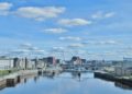 Cityscape of Glasgow with modern buildings lining both sides of the River Clyde under a bright blue sky scattered with fluffy clouds; a calm and serene atmosphere.