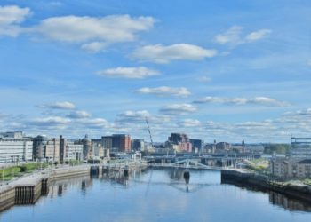 Cityscape of Glasgow with modern buildings lining both sides of the River Clyde under a bright blue sky scattered with fluffy clouds; a calm and serene atmosphere.
