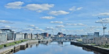 Cityscape of Glasgow with modern buildings lining both sides of the River Clyde under a bright blue sky scattered with fluffy clouds; a calm and serene atmosphere.