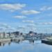 Cityscape of Glasgow with modern buildings lining both sides of the River Clyde under a bright blue sky scattered with fluffy clouds; a calm and serene atmosphere.