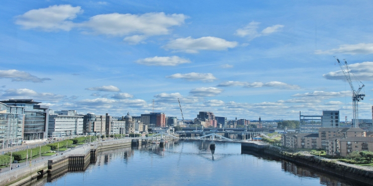 Cityscape of Glasgow with modern buildings lining both sides of the River Clyde under a bright blue sky scattered with fluffy clouds; a calm and serene atmosphere.