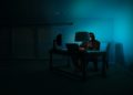 Person sits in a dimly lit room, illuminated by a computer screen and a lamp, conveying a mysterious and secretive tone.