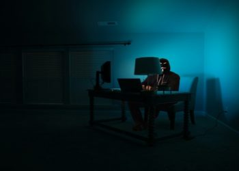 Person sits in a dimly lit room, illuminated by a computer screen and a lamp, conveying a mysterious and secretive tone.