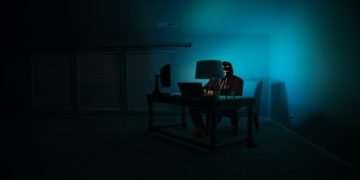 Person sits in a dimly lit room, illuminated by a computer screen and a lamp, conveying a mysterious and secretive tone.