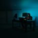 Person sits in a dimly lit room, illuminated by a computer screen and a lamp, conveying a mysterious and secretive tone.
