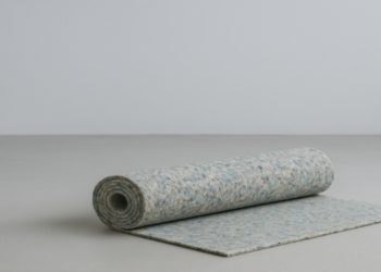 Rolled-up, speckled blue and gray carpet rests on a light gray surface with one edge unfurled. The minimalist setting conveys calm and simplicity.