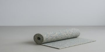Rolled-up, speckled blue and gray carpet rests on a light gray surface with one edge unfurled. The minimalist setting conveys calm and simplicity.