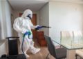 Person in a full protective suit and mask uses a spray device to disinfect a bright, modern dining room. The mood is serious and focused.