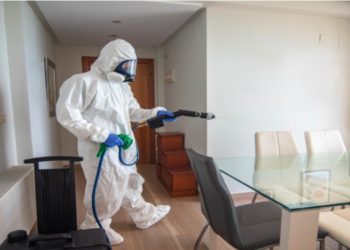 Person in a full protective suit and mask uses a spray device to disinfect a bright, modern dining room. The mood is serious and focused.