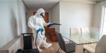 Person in a full protective suit and mask uses a spray device to disinfect a bright, modern dining room. The mood is serious and focused.