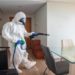 Person in a full protective suit and mask uses a spray device to disinfect a bright, modern dining room. The mood is serious and focused.