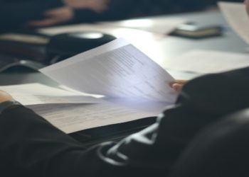 Person in a suit holds business papers at a conference table, surrounded by documents and a notebook. The setting conveys a focused, professional atmosphere.