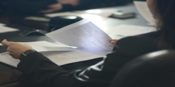 Person in a suit holds business papers at a conference table, surrounded by documents and a notebook. The setting conveys a focused, professional atmosphere.