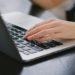 Hand typing on a laptop placed on a dark wooden table. The image conveys productivity and focus, with a close-up view of the keyboard.