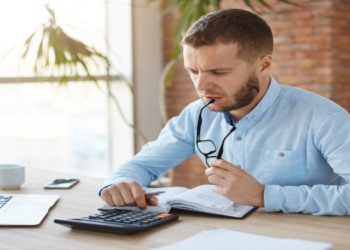 Man in a blue shirt sits at a desk with a laptop, cup, and phone, focusing intently on a calculator. He appears thoughtful, with glasses in hand.