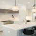 Modern kitchen with white cabinetry, a central island, black chairs, and pendant lights. The space exudes elegance and brightness, with marble accents.