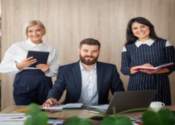 Three professionals pose cheerfully in an office setting. A woman on the left holds a tablet, a man in the center sits at a desk with a laptop, and a woman on the right holds a book.
