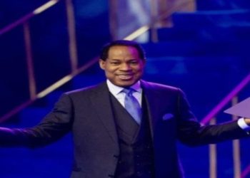 Pastor Chris Oyakhilome in a suit stands on a stage with open arms, holding a book. The lighting is blue and purple, creating an energetic and welcoming atmosphere.