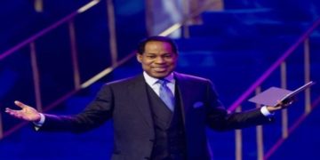 Pastor Chris Oyakhilome in a suit stands on a stage with open arms, holding a book. The lighting is blue and purple, creating an energetic and welcoming atmosphere.