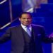 Pastor Chris Oyakhilome in a suit stands on a stage with open arms, holding a book. The lighting is blue and purple, creating an energetic and welcoming atmosphere.