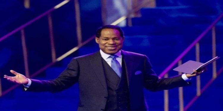 Pastor Chris Oyakhilome in a suit stands on a stage with open arms, holding a book. The lighting is blue and purple, creating an energetic and welcoming atmosphere.