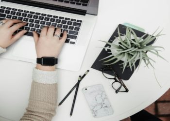 Hands typing on a laptop on a round white table, accompanied by a plant, glasses, pencils, and a marble-patterned phone, conveying productivity.