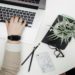 Hands typing on a laptop on a round white table, accompanied by a plant, glasses, pencils, and a marble-patterned phone, conveying productivity.