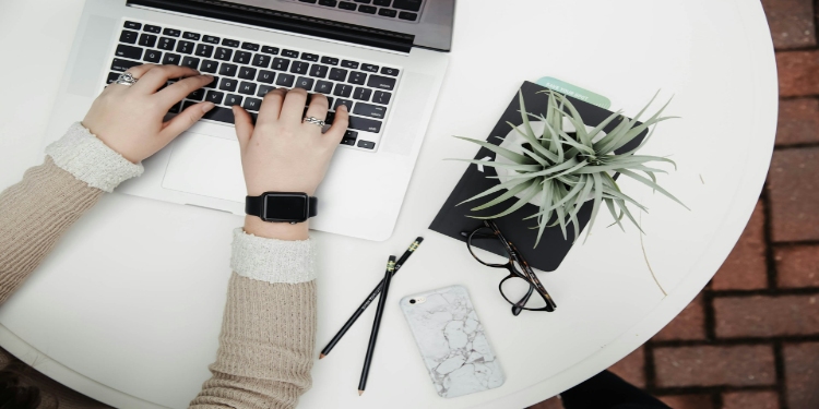 Hands typing on a laptop on a round white table, accompanied by a plant, glasses, pencils, and a marble-patterned phone, conveying productivity.