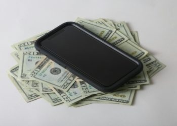 A smartphone rests on a pile of U.S. twenty-dollar bills against a neutral background, conveying wealth or technology expense.