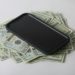 A smartphone rests on a pile of U.S. twenty-dollar bills against a neutral background, conveying wealth or technology expense.
