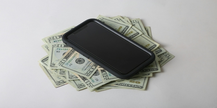 A smartphone rests on a pile of U.S. twenty-dollar bills against a neutral background, conveying wealth or technology expense.