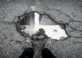 Aerial view of a sidewalk pothole reflecting skyscrapers and a flying airplane, with a person’s shoes visible at the bottom edge, conveying urban life.
