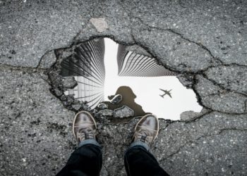Aerial view of a sidewalk pothole reflecting skyscrapers and a flying airplane, with a person’s shoes visible at the bottom edge, conveying urban life.