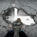 Aerial view of a sidewalk pothole reflecting skyscrapers and a flying airplane, with a person’s shoes visible at the bottom edge, conveying urban life.