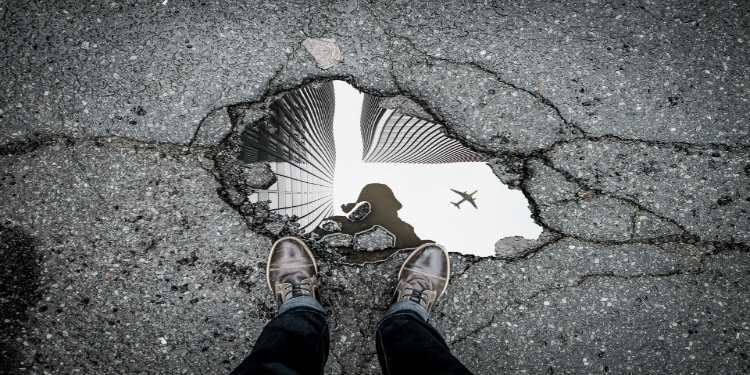 Aerial view of a sidewalk pothole reflecting skyscrapers and a flying airplane, with a person’s shoes visible at the bottom edge, conveying urban life.