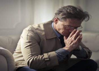 Middle-aged man in a beige coat sits on a couch, hands pressed together in thought, conveying a contemplative and introspective mood.