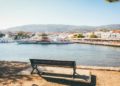 Peaceful waterfront scene with a wooden bench overlooking Skiathos, Greece filled with boats. Sunlit trees frame the view, and quaint buildings line the shore.
