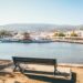 Peaceful waterfront scene with a wooden bench overlooking Skiathos, Greece filled with boats. Sunlit trees frame the view, and quaint buildings line the shore.