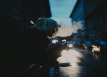 Person with short blonde hair smokes in a dimly lit street at dusk. Car headlights illuminate the scene, creating a moody, contemplative atmosphere.