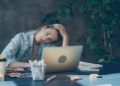 Woman in a denim jacket sits at a desk, looking fatigued. Her hand rests on her head, surrounded by a laptop, coffee cup, and plants, conveying exhaustion.