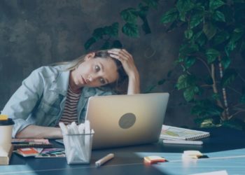 Woman in a denim jacket sits at a desk, looking fatigued. Her hand rests on her head, surrounded by a laptop, coffee cup, and plants, conveying exhaustion.