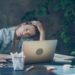 Woman in a denim jacket sits at a desk, looking fatigued. Her hand rests on her head, surrounded by a laptop, coffee cup, and plants, conveying exhaustion.