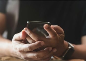 erson wearing a black shirt holds a smartphone with both hands, focusing intently. The casual setting conveys an engaged, thoughtful mood.