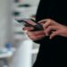 Person using a smartphone indoors. Their hands focus on typing, with a blurred office desk in the background, conveying a tech-savvy atmosphere.