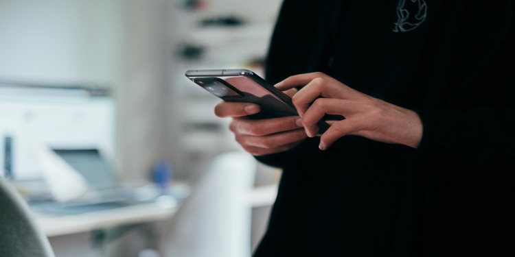 Person using a smartphone indoors. Their hands focus on typing, with a blurred office desk in the background, conveying a tech-savvy atmosphere.