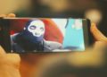 Hands holding a smartphone displaying a masked character with a skull design on the mask. The background is blurred, creating a focused, intense scene.