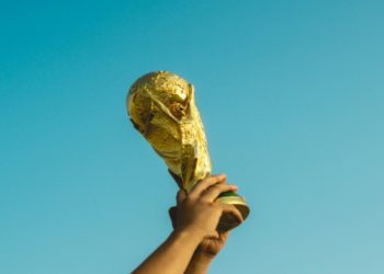Hands raise a golden trophy against a clear blue sky, symbolizing triumph and celebration. The moment is uplifting and victorious.
