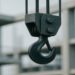 Close-up of a black crane hook suspended by cables, with a blurred background of an unfinished concrete building, conveying an industrial tone.