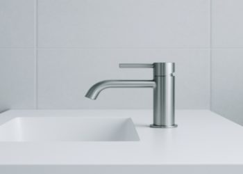 Sleek modern bathroom with a minimalist design. A metallic faucet extends over a white countertop with an inset sink, set against light gray tiled walls.